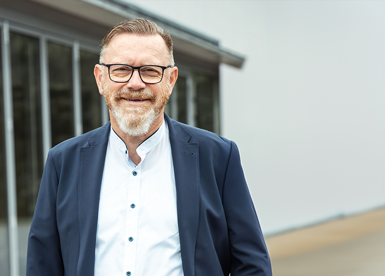 Ein lächelnder Mann mit Brille, Bart und kurzen Haaren steht draußen vor einem modernen Gebäude. Er trägt einen blauen Blazer über einem weißen Hemd mit Knöpfen und strahlt damit ein professionelles Erscheinungsbild aus.