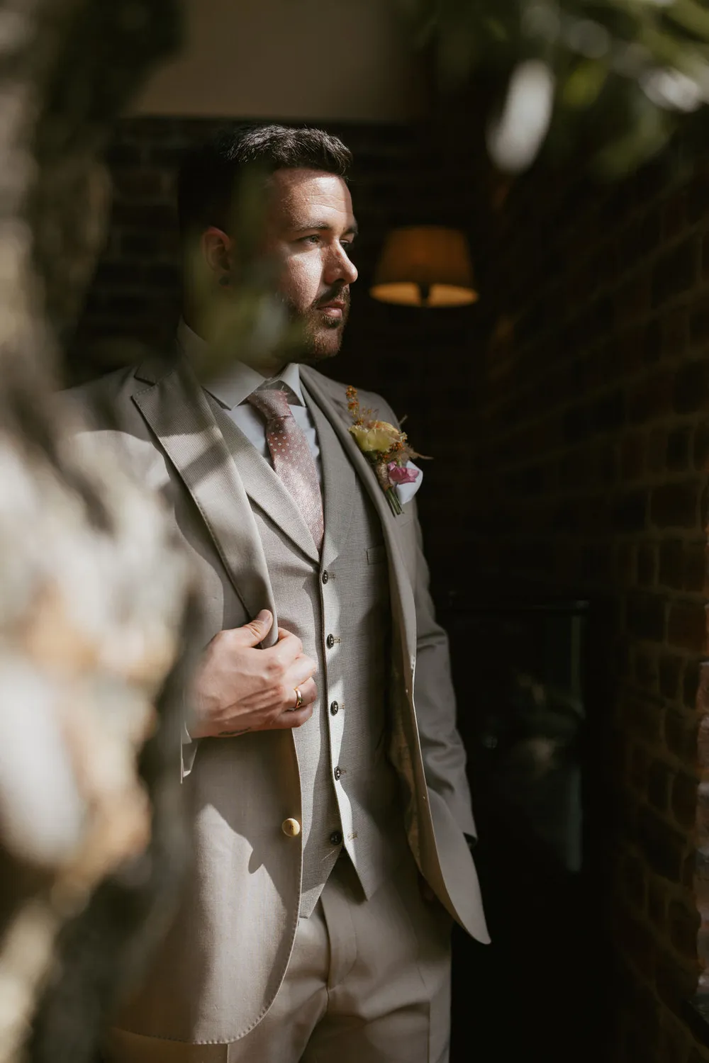 Ein Mann in einem dreiteiligen beigen Anzug mit rosa Krawatte und Blumenboutonniere steht nachdenklich neben einer Backsteinmauer. Weiches, warmes Licht hebt sein Profil hervor und im Vordergrund ist teilweise Grün zu sehen.