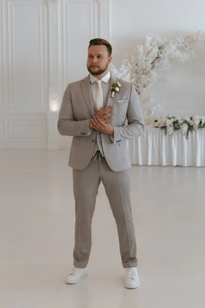 Eine Person in hellgrauem Anzug und weißen Turnschuhen steht in einem hellen, geräumigen Raum mit elegantem weißen Dekor und einem Blumenarrangement im Hintergrund. Sie trägt eine Boutonniere am Revers und faltet die Hände.