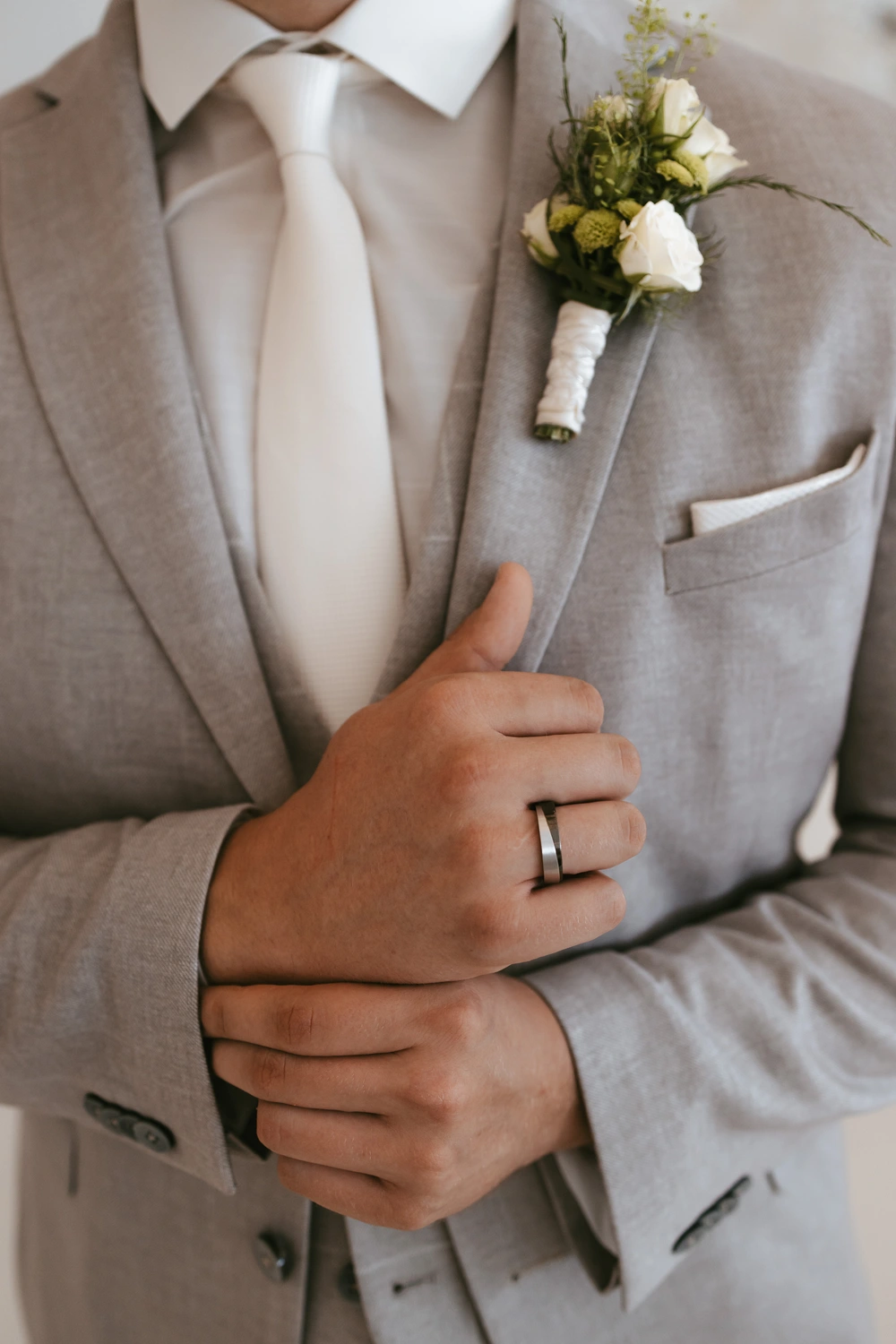 Ein Mann in einem hellgrauen Anzug mit weißem Hemd und Krawatte hält seine Hände zusammen. An seiner linken Hand trägt er einen silbernen Ring. An seinem Revers ist eine Ansteckblume mit weißen Blumen und Grünpflanzen befestigt.