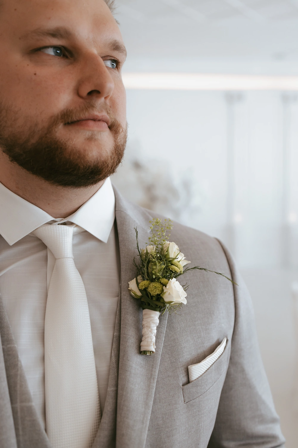 Ein Mann in hellgrauem Anzug mit weißem Hemd und Krawatte, am Revers trägt er eine Ansteckblume aus weißen Blumen und Grünpflanzen, blickt in die Ferne. Der Hintergrund ist leicht unscharf.