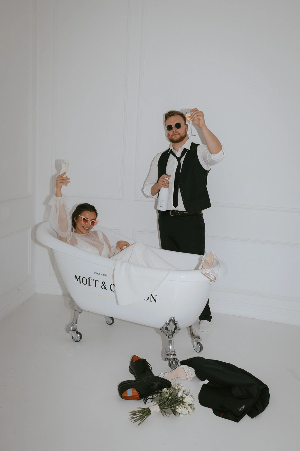 Eine Frau in einem weißen Kleid und mit roter Sonnenbrille sitzt in einer Badewanne und hält ein Champagnerglas in der Hand. Neben ihr steht ein Mann in Anzug und Sonnenbrille, der ebenfalls ein Glas hält. Auf dem Boden liegen Blumen und eine Jacke. Beide scheinen zu feiern.