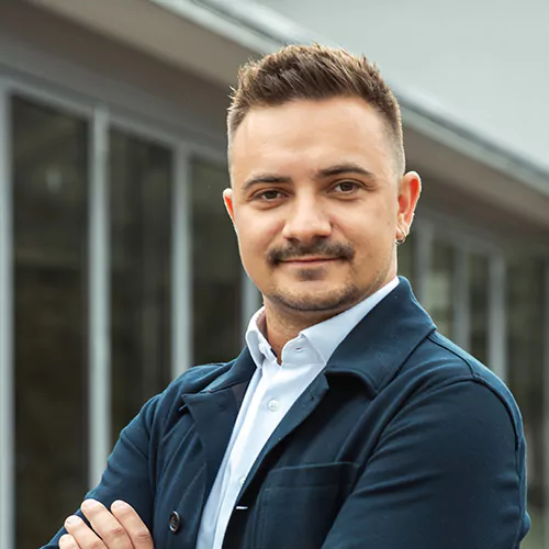 Eine Person mit kurzen Haaren steht selbstbewusst vor einem Gebäude mit großen Fenstern. Sie trägt eine dunkle Jacke und ein weißes Hemd. Sie lächelt leicht und hat die Arme verschränkt.