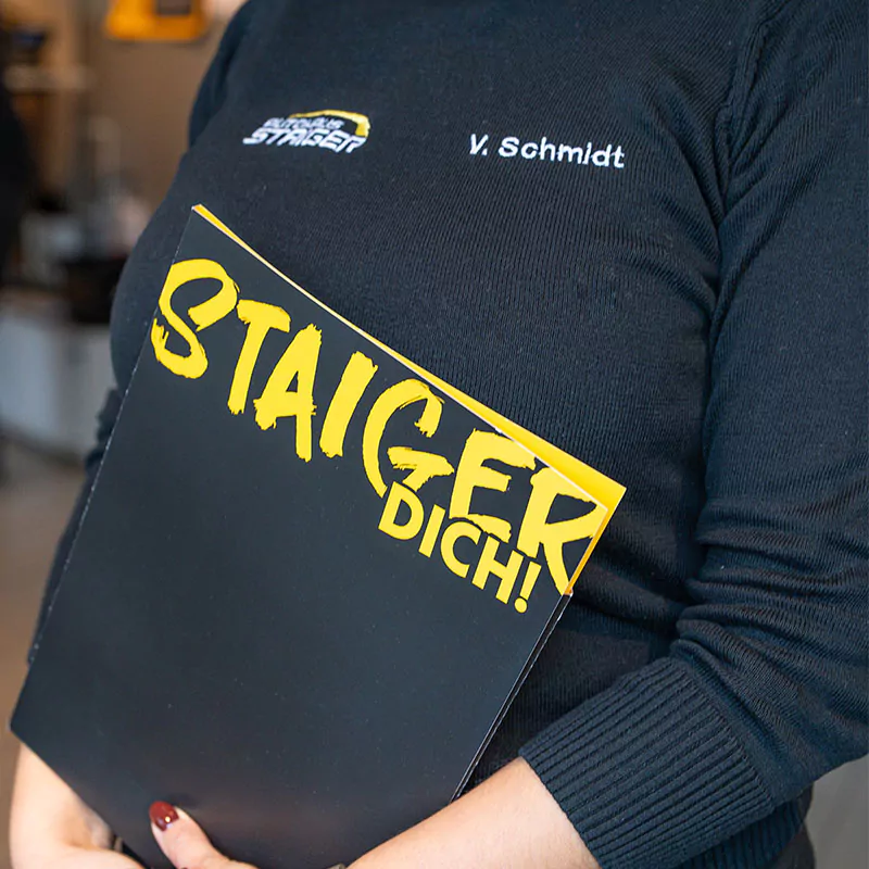 Eine Person hält einen schwarz-gelben Ordner mit dem Text STAIGER DICH! Die Person trägt ein schwarzes Shirt mit einem auf der Brust aufgestickten Namen.