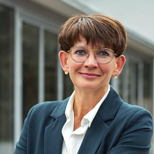 Eine Person mit kurzen braunen Haaren, Brille, dunklem Blazer und weißem Hemd, lächelt leicht. Im Hintergrund ist die verschwommene Außenansicht eines Gebäudes mit großen Fenstern zu sehen.