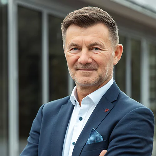 Ein Mann in einem marineblauen Blazer steht mit verschränkten Armen im Freien und lächelt leicht. Er hat kurze Haare und einen gestutzten Bart. Im Hintergrund sind die Glasfenster eines Gebäudes zu sehen.