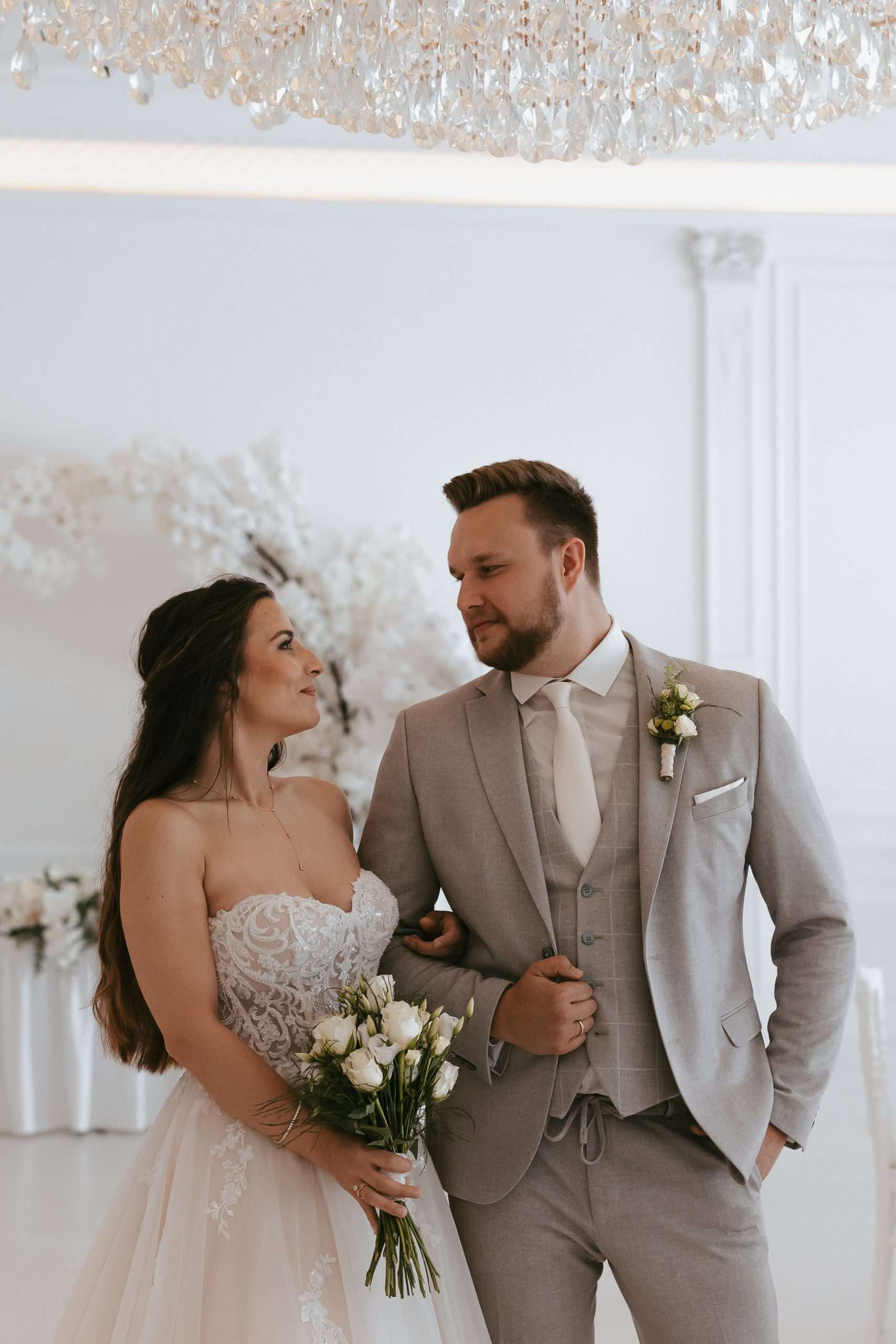 Ein Mann und eine Frau vor ihrer Hochzeit. Die Frau trägt einen weißes mit Blumen verziertes Kleid sowie zusätzlich einem Blumenstrauß. Der Mann ist in einem beigen Anzug mit weißem Hemd und weißer Krawatte mit einem Ehering