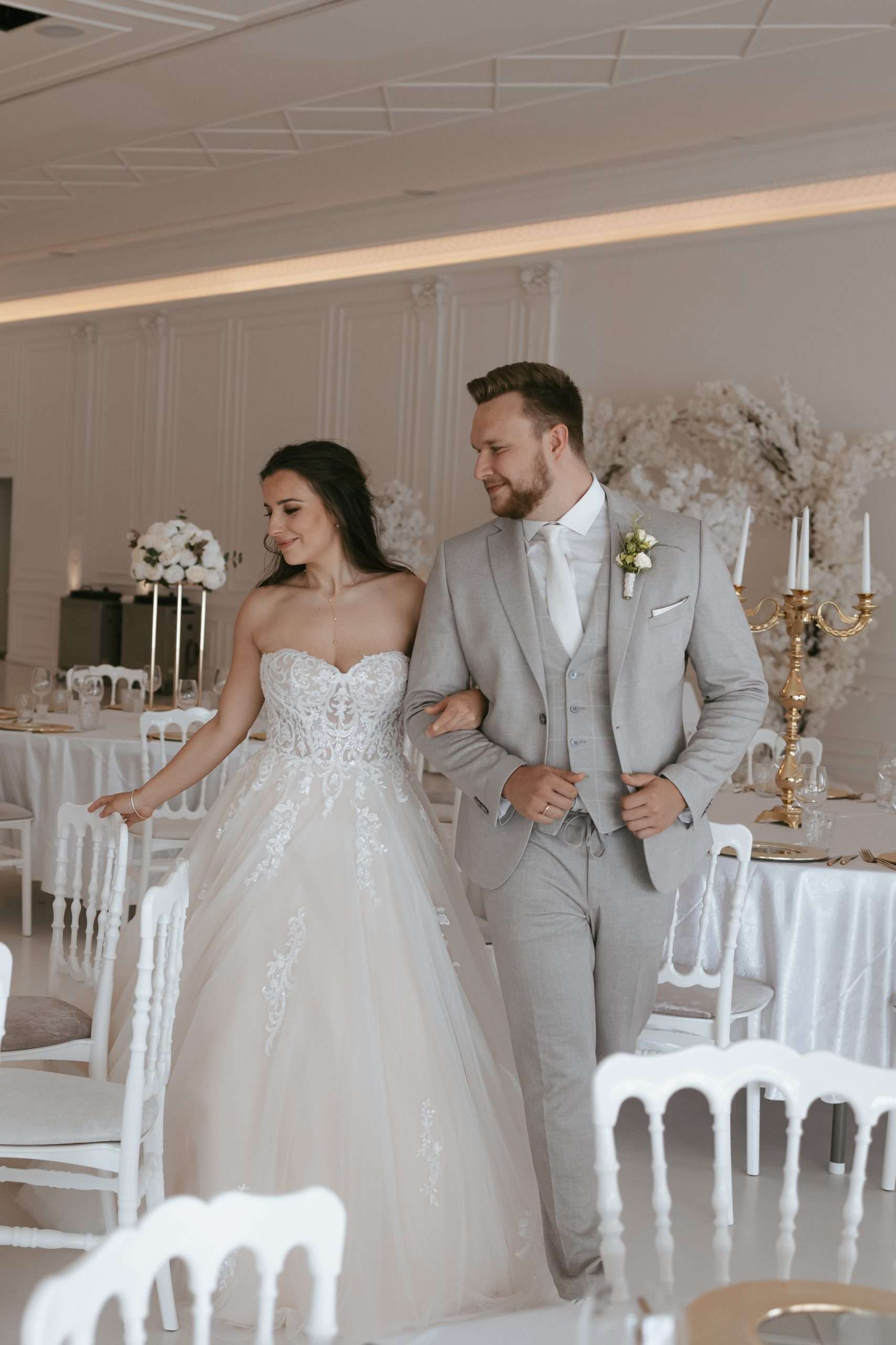 Braut und Bräutigam stehen zusammen in formeller Kleidung. Der Bräutigam trägt einen schwarzen Anzug mit einer Ansteckblume und die Braut trägt ein weißes Kleid und hält einen Strauß weißer Blumen. Im Hintergrund sind weiße Blumendekorationen zu sehen.