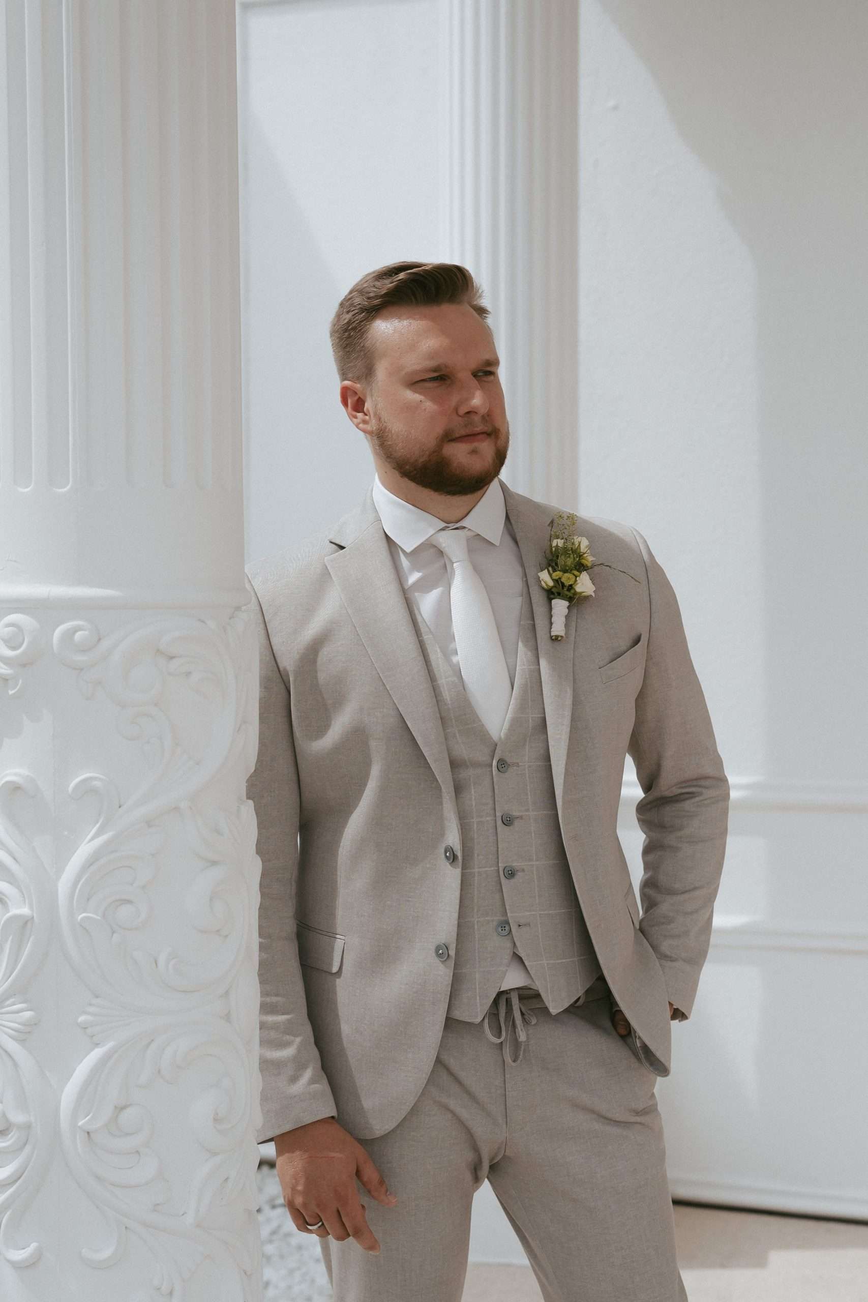 Draußen auf einer Hochzeit mit verzierten Säulen steht ein Mann in einem beigen Anzug mit weißem Hemd und weißer Krawatte mit einem Ehering