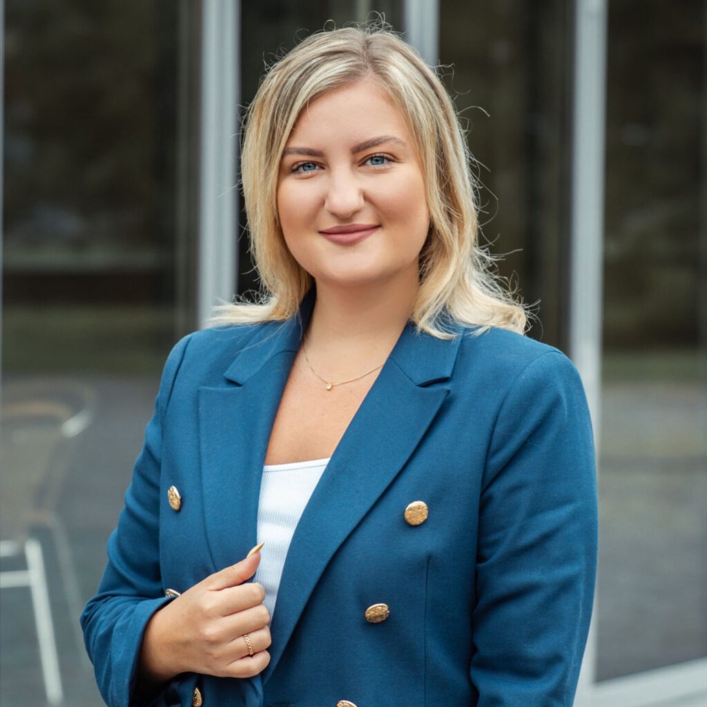 Eine blonde Frau in einem blauen Anzug und Jeans mit weißem Shirt steht draußen vor einer unscharfen Glaswand.