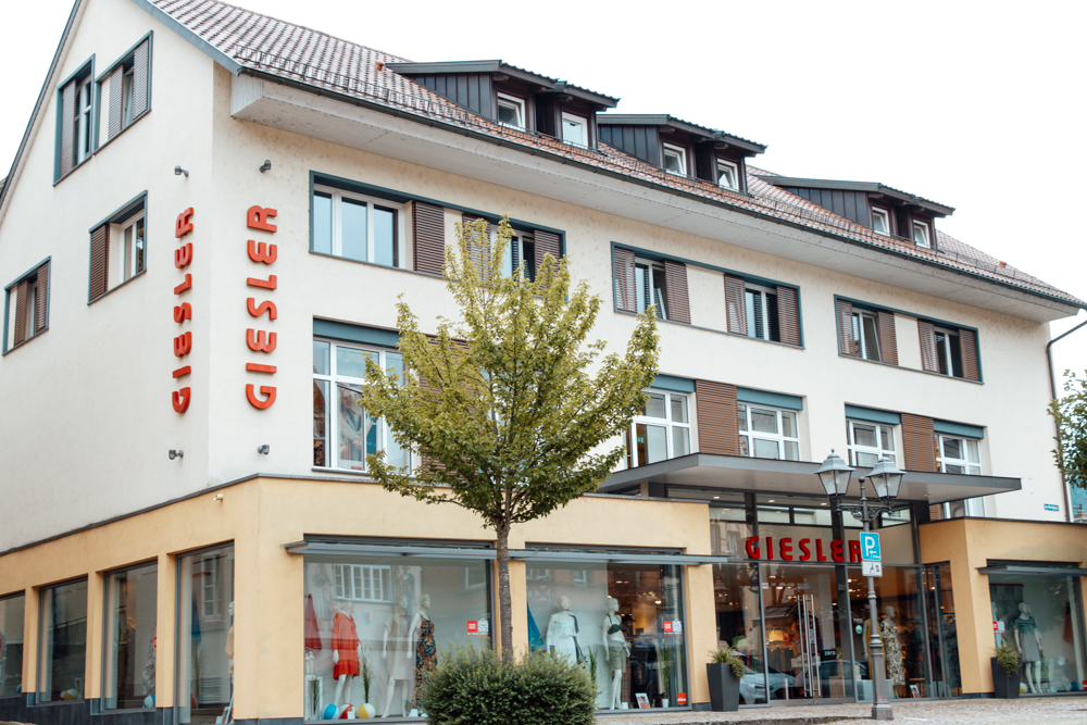 Ein creme farbenes Gebäude mit Spitzdach und der Aufschrift GIESLER sowie gekleideten Puppen im Schaufenster.