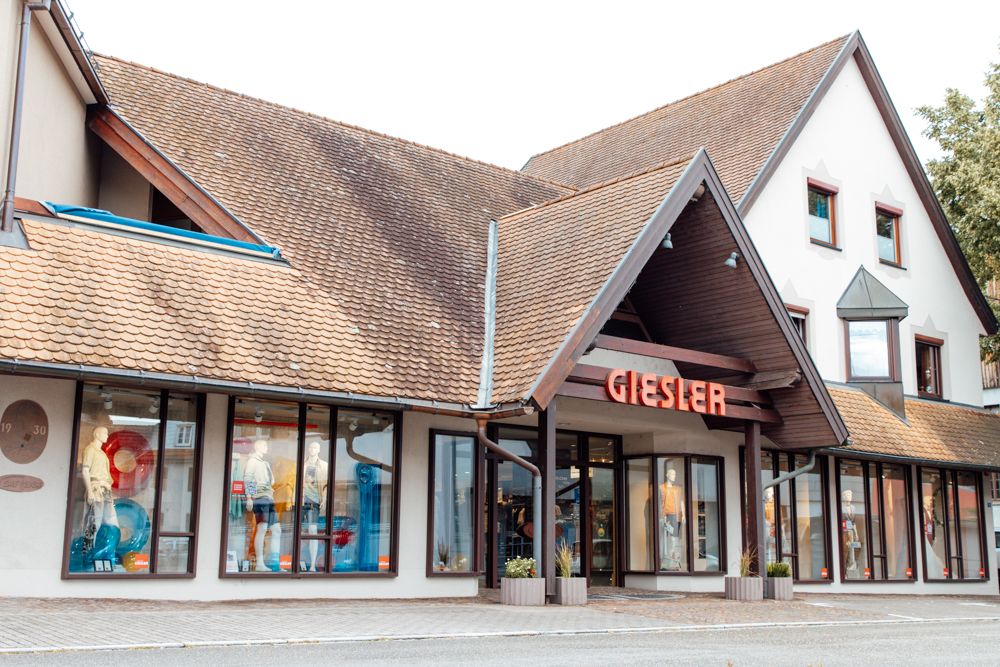 Ein weißes Gebäude mit Holzbalken und Spitzdach mit der Aufschrift GIESLER mit gekleideten Puppen im Schaufenster.