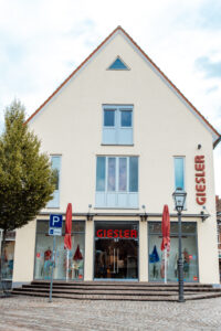 Ein creme farbenes Gebäude mit rotem Spitzdach und der Aufschrift GIESLER mit gekleideten Puppen im Schaufenster und zwei roten Schirmen vor dem Eingang.