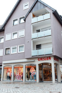 Ein graues Gebäude mit Spitzdach und der Aufschrift GIESLER trend sowie gekleideten Puppen im Schaufenster.