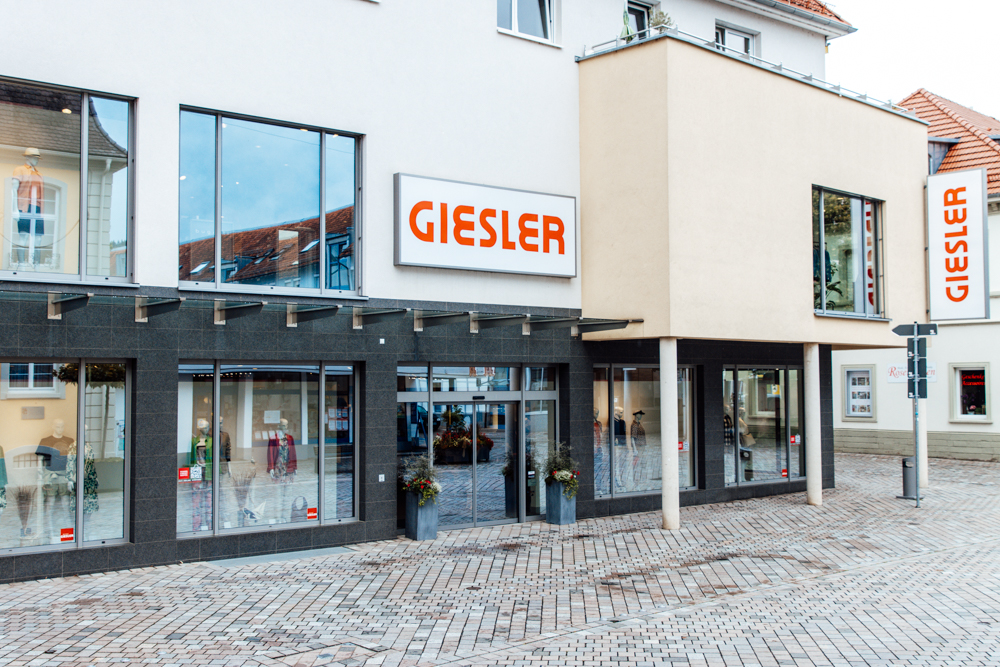 Ein creme farbenes Gebäude mit der Aufschrift GIESLER sowie gekleideten Puppen im Schaufenster.