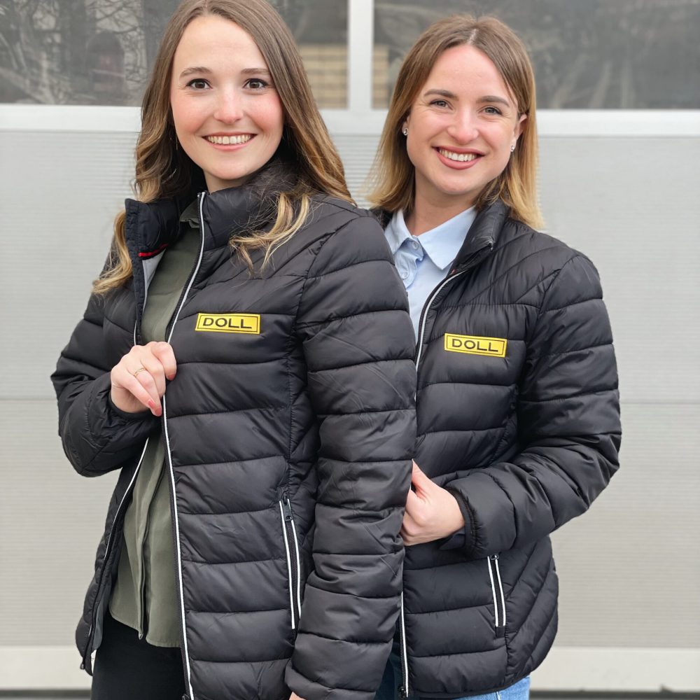 Zwei Frauen stehen vor einer Garage. Beide haben eine schwarze Steppjacke mit der Aufschrift DOLL an.