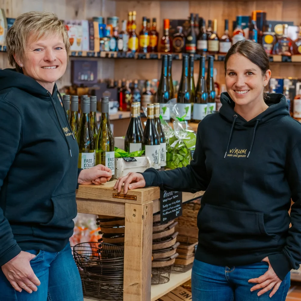 Zwei lächelnde Menschen in Kapuzenpullovern stehen in einem Laden voller Flaschen und verschiedener Produkte. Sie posieren neben einer Holzvitrine mit Weinflaschen. In den Regalen im Hintergrund finden sich verschiedene alkoholische Getränke.