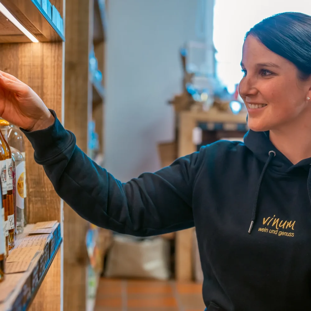 Eine Frau mit einem schwarzen Kapuzenpullover mit der Aufschrift „Vinum“ lächelt und greift nach einer Flasche auf einem Holzregal in einem Geschäft. Um sie herum sind Flaschen in den Regalen aufgereiht. Der Laden ist innen sanft beleuchtet.