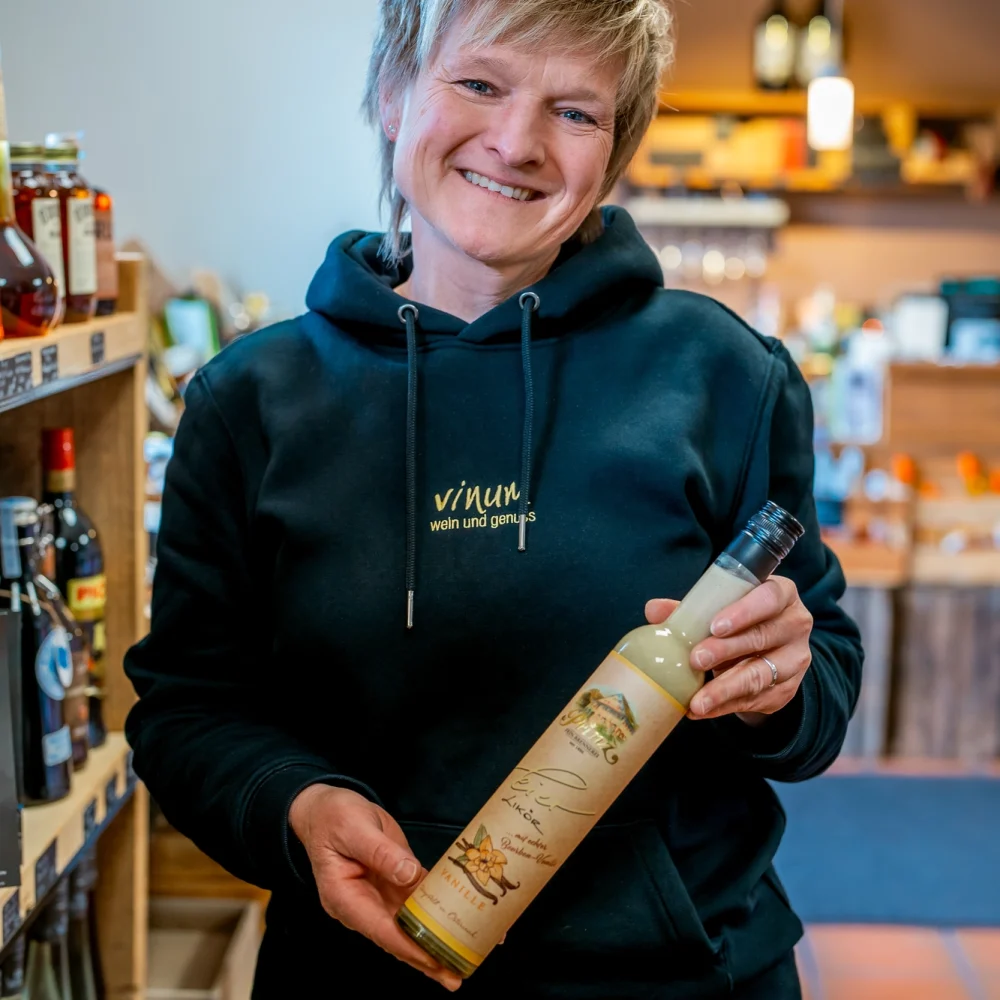 Eine Person in einem schwarzen Kapuzenpullover lächelt, während sie in einem Geschäft eine Flasche hält. Die Regale dahinter sind mit verschiedenen Flaschen gefüllt und die warme Beleuchtung sorgt für eine gemütliche Atmosphäre.