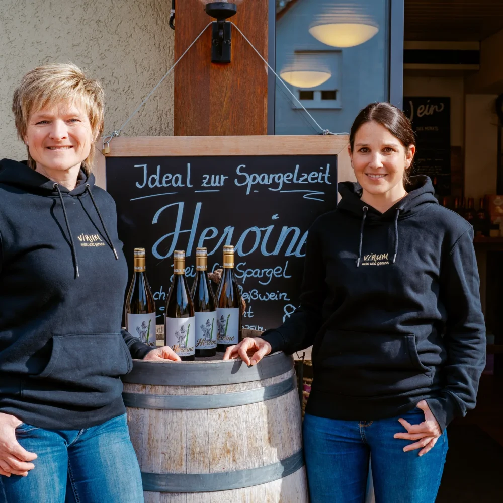 Zwei Frauen in schwarzen Kapuzenpullovern stehen neben einem Holzfass, in dem Weinflaschen ausgestellt sind, vor einem Geschäft. Auf einer Tafel hinter ihnen steht „Ideal zur Spargelzeit und Heppni“. Die Atmosphäre ist leger und einladend.