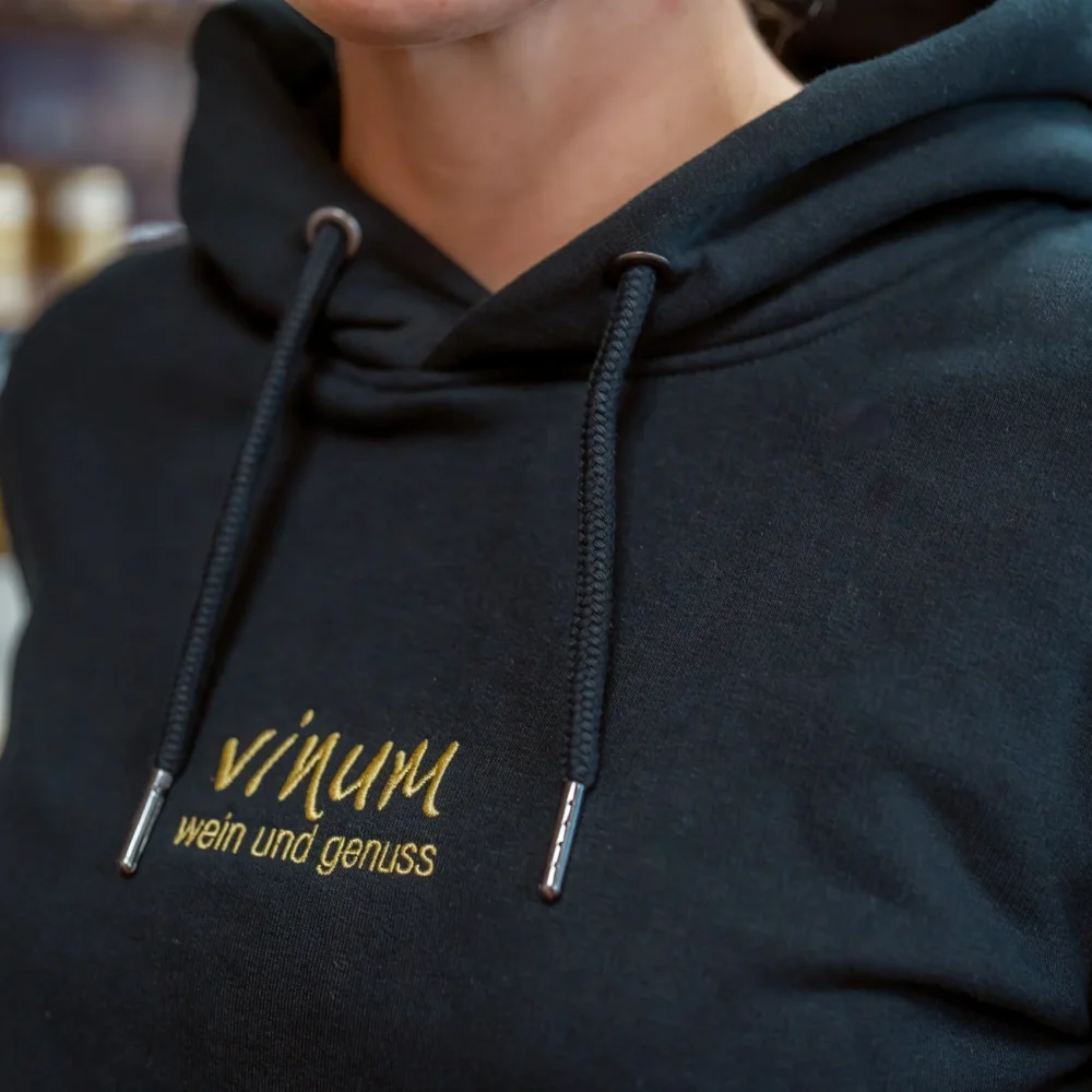 Nahaufnahme einer Person, die einen dunklen Kapuzenpullover mit gelbem Text trägt, auf dem „vinum wein und genuss“ steht. Weinflaschen sind im Hintergrund verschwommen.