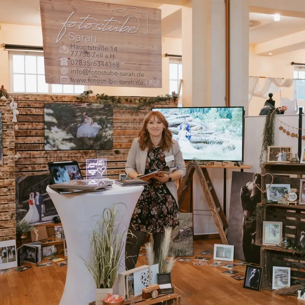 Eine Frau steht an einem Stand mit Fotoausrüstung und Bildern. Der Aufbau umfasst einen Hintergrund mit dem Logo der Fotostube Sarah und einen Fernseher, auf dem eine Diashow läuft. Der Stand ist mit Fotos, Holzkisten und einem kleinen Tisch dekoriert.