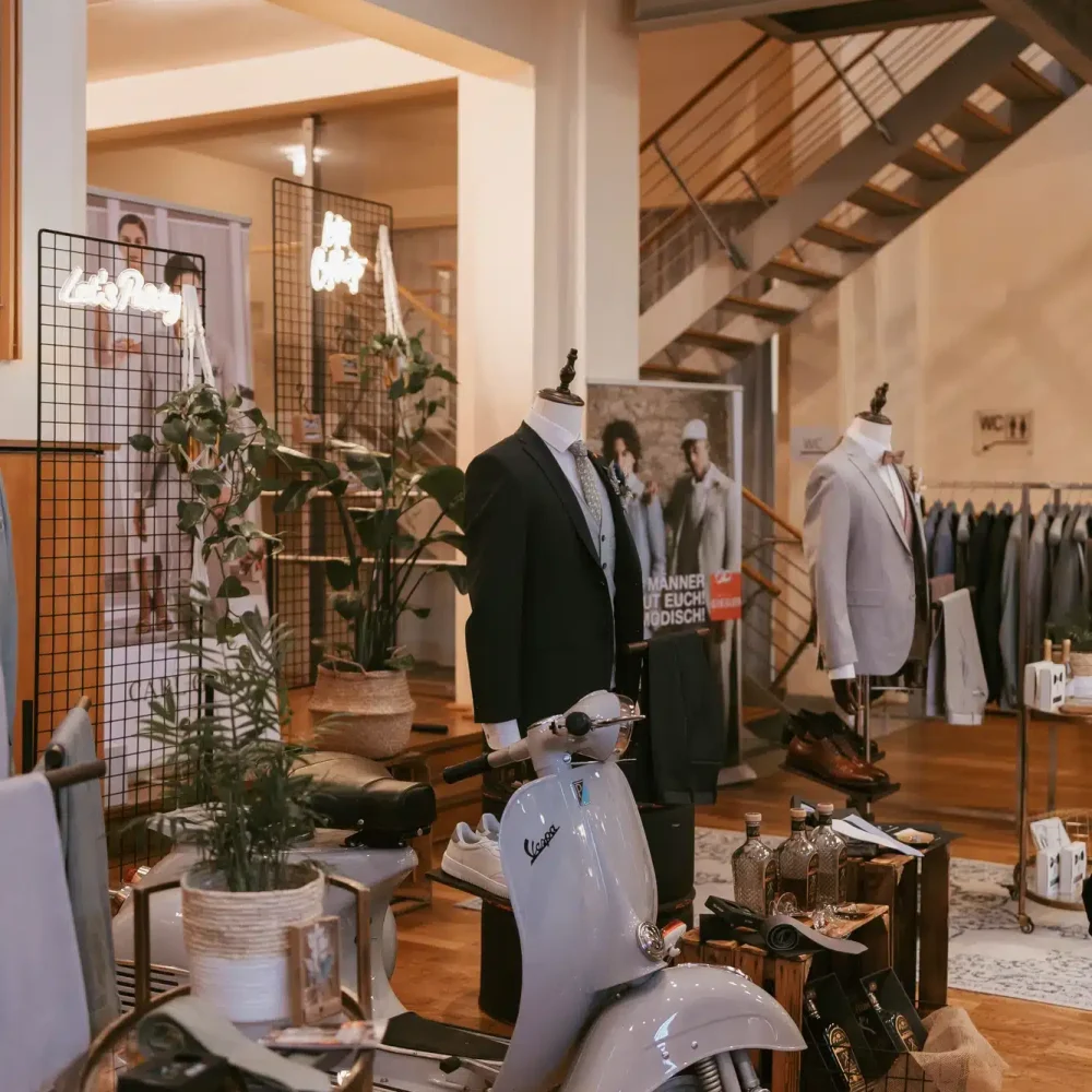 In einem schicken Herrenbekleidungsgeschäft hängen Anzüge an Schaufensterpuppen. In der Mitte steht ein Oldtimer-Roller, umgeben von Pflanzen und Koffern. Im Hintergrund sind eine Treppe und Kleiderständer zu sehen, die eine moderne, schicke Atmosphäre schaffen.