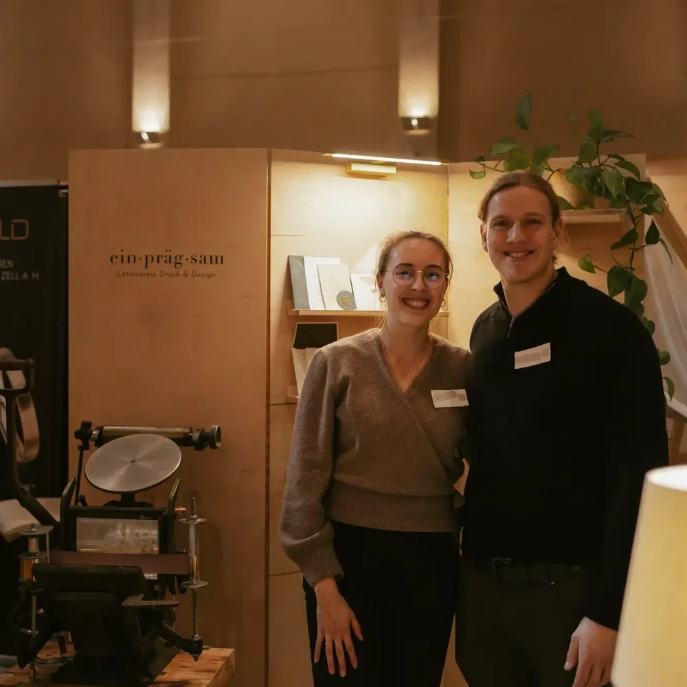 Zwei lächelnde Menschen stehen an einem Stand, an dem Druckgeräte und Papierwaren angeboten werden. Im Hintergrund ist ein Schild mit der Aufschrift „ein präg:sam“ zu sehen. Der Stand ist warm beleuchtet und verfügt über hohe Decken und sichtbare Fenster.