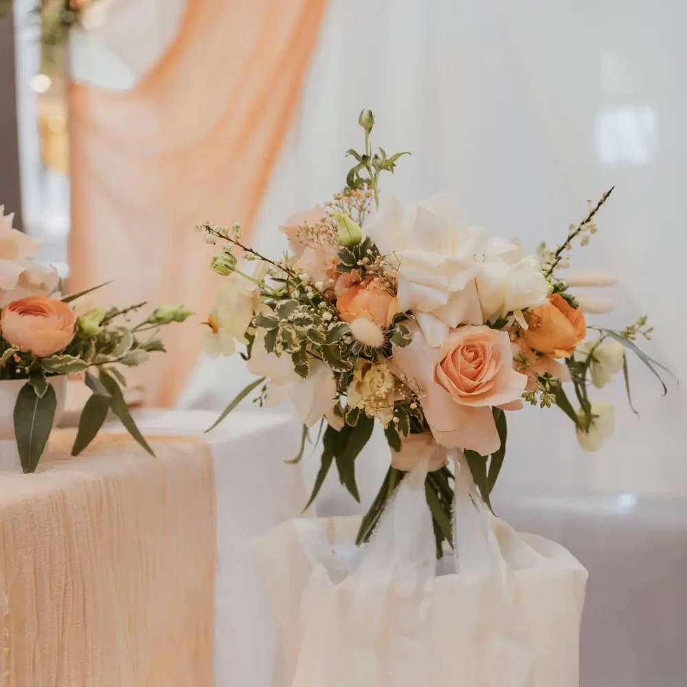 Ein Blumenarrangement aus pfirsichfarbenen und weißen Rosen mit Grünpflanzen, platziert auf einem weiß drapierten Ständer. Ein passender Blumenstrauß schmückt einen Tisch in der Nähe, vor einem pfirsichfarbenen Stoffhintergrund. Die Szene strahlt Eleganz und ein sanftes, romantisches Ambiente aus.