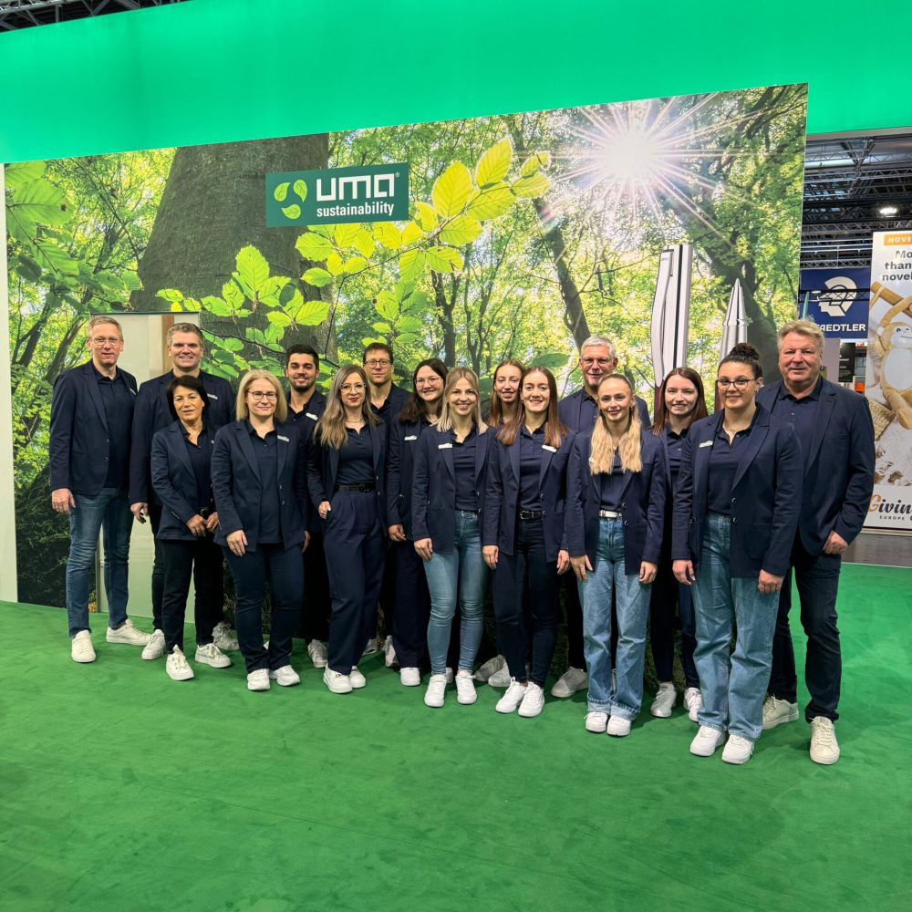 Viele Personen stehen im dunkelblauen Anzug, dunkelblauen Hemd und weißen Schuhen in einer Messehalle vor einem grünen Hintergrund mit einem Banner der die Natur zeigt und das Logo mit dem Namen UMA.