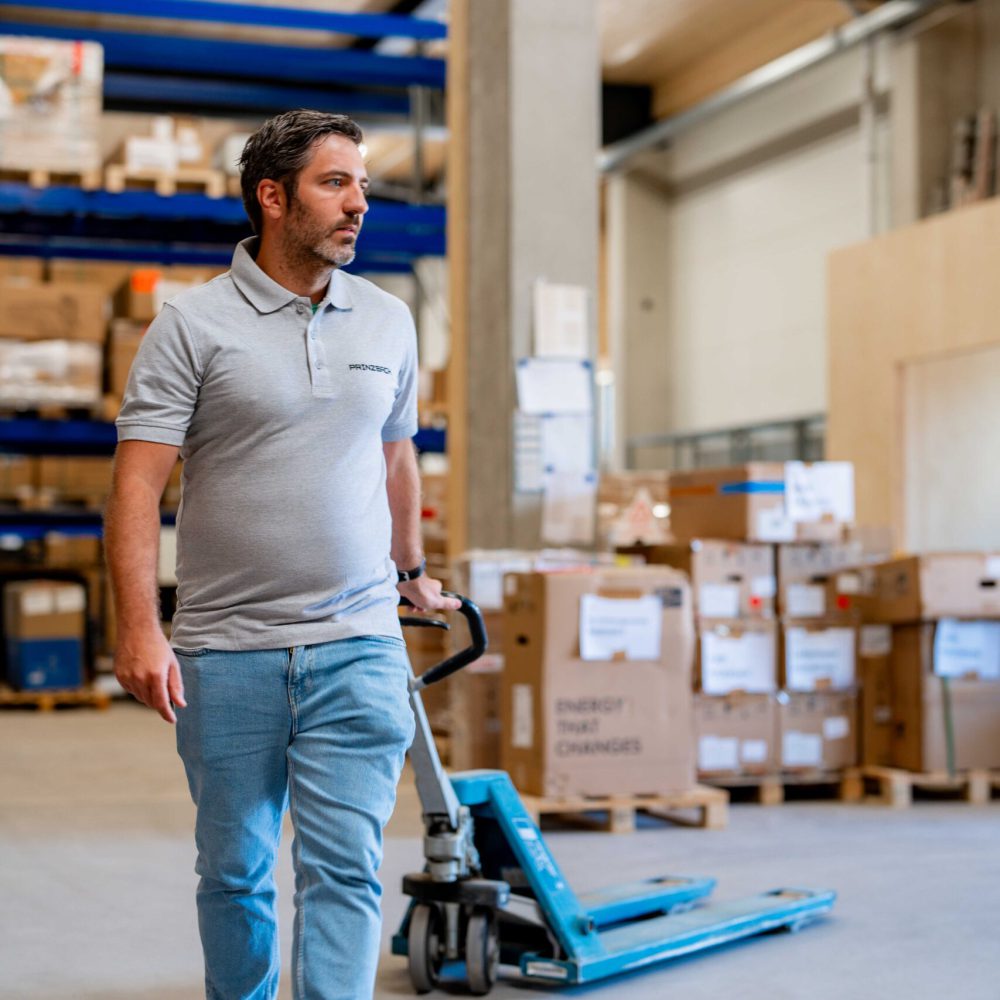 Ein Mann mit grauem Polo-Shirt und der Aufschrift PRINZBACH sowie blauer Jeans befindet sich in einem Lagerraum.