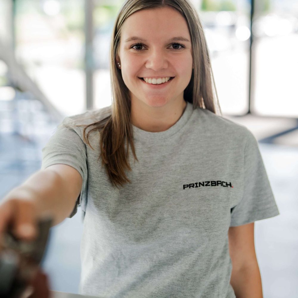 Eine Frau trägt ein hellgraues Shirt mit dem schwarz aufgestickten Wort PRINZBACH trägt. Die Frau im Shirt steht vor einem unscharfen Hintergrund.