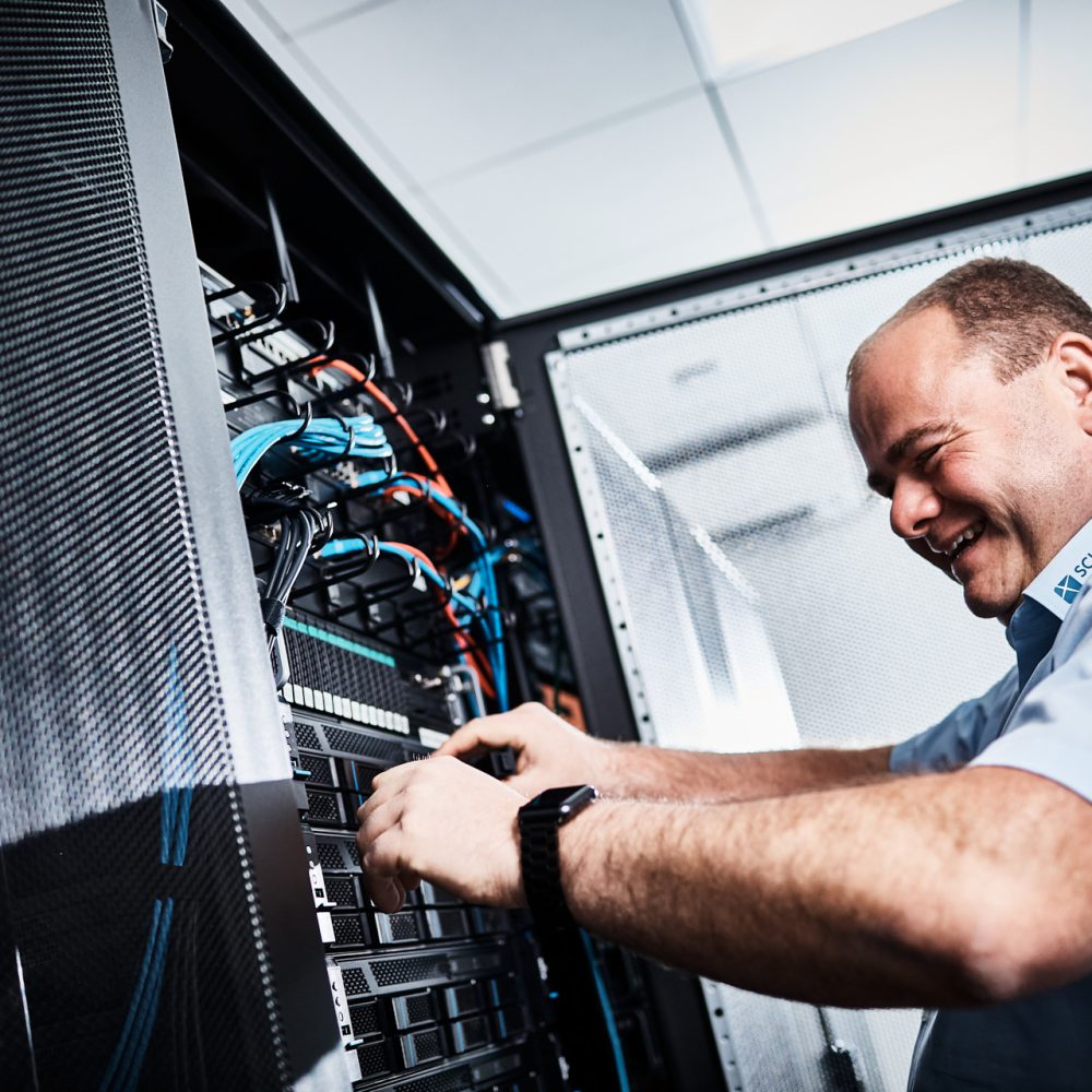 Ein Mann im hellblauen Hemd arbeitet an einem Server. Dabei lacht er und trägt eine Smartwatch.