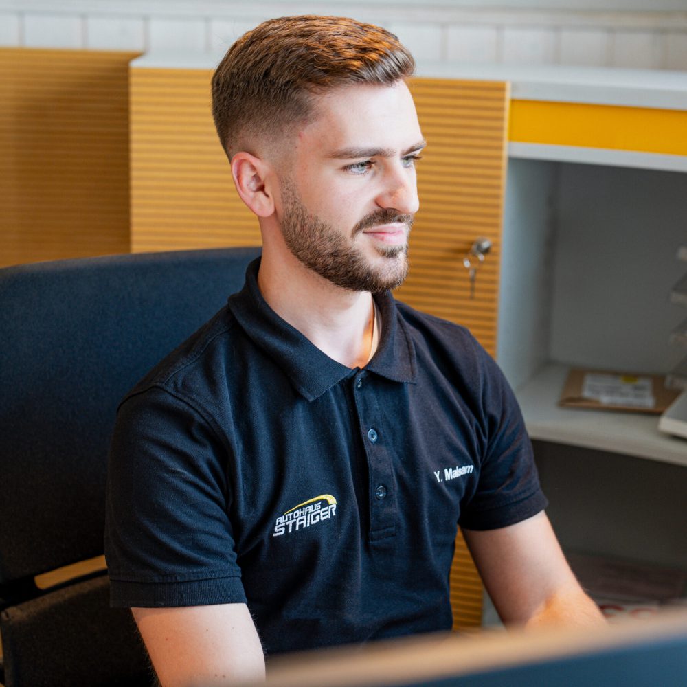 Ein junger Mann sitzt in einem Bürogebäude auf einem schwarzen Bürostuhl mit einem schwarzen Polo und der Aufschrift Autohaus Staiger