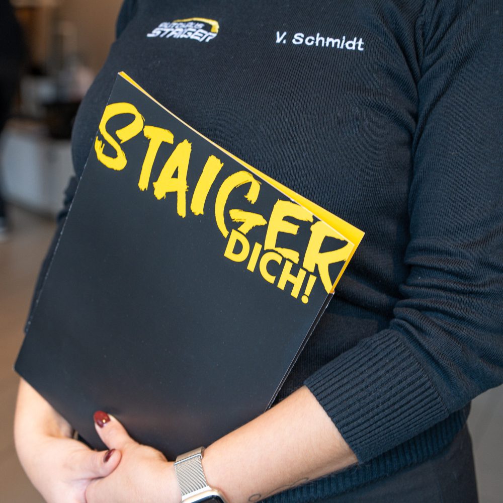 Eine blonde Frau mit schwarzen Pullover mit der Aufschrift Autohaus Staiger hält eine schwarze Mappe in ihren Händen mit der Aufschrift STAIGER DICH!