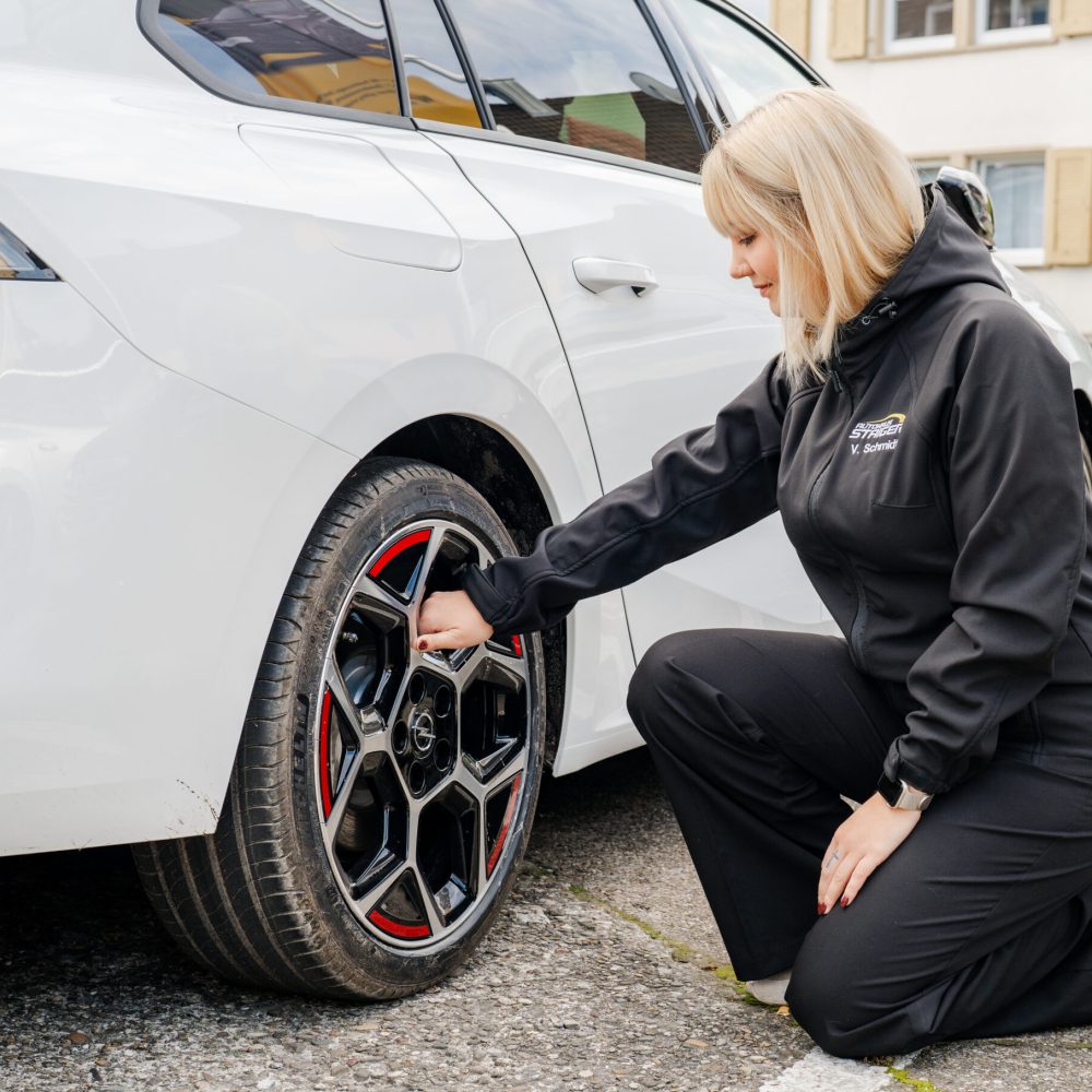 Eine blonde Frau mit schwarzer Jacke und der Aufschrift Autohaus Staiger kniet draußen vor einem weißen Auto der Marke Opel und hält die Felge des Autos fest